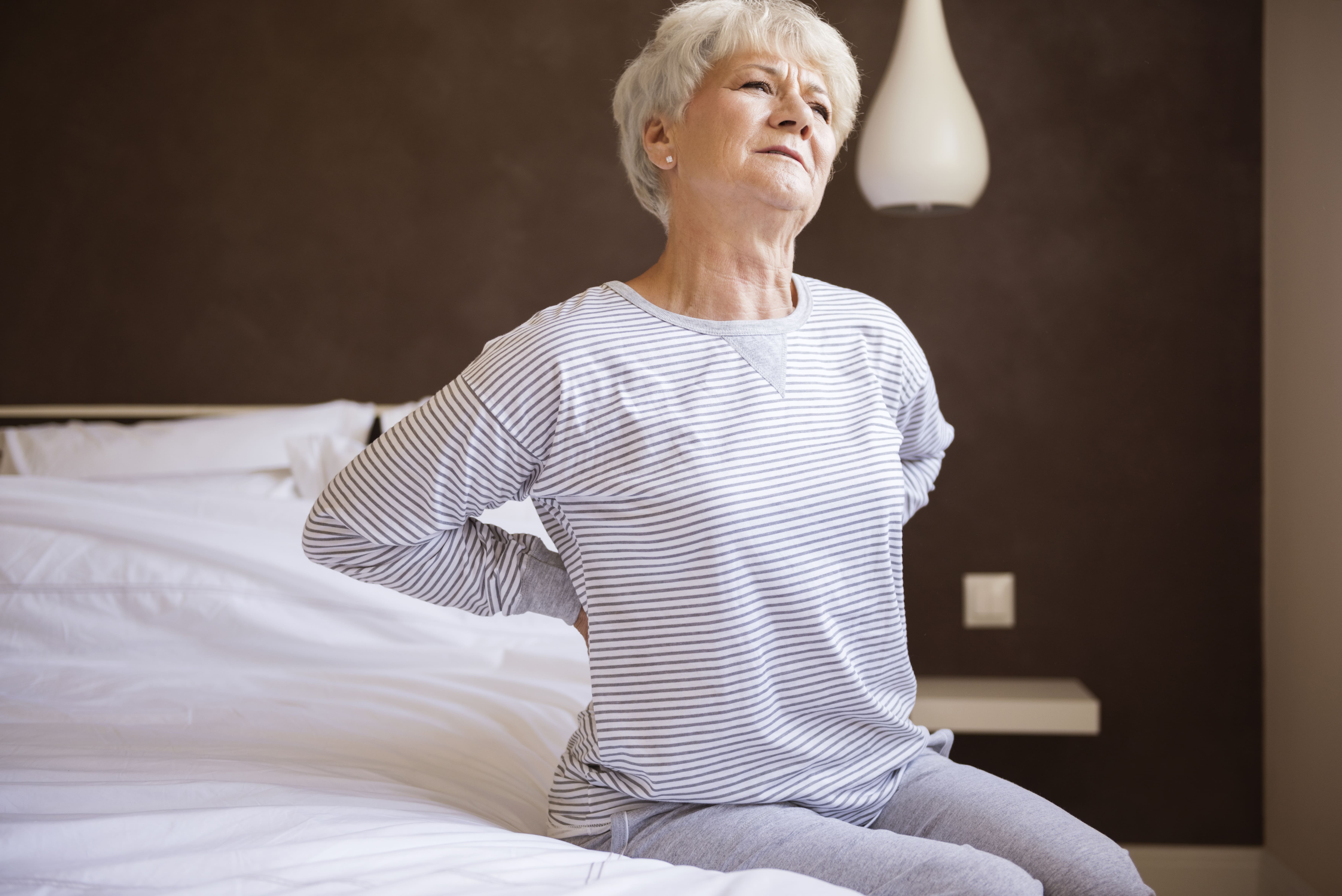 An older woman struggling with back pain as she gets up from bed ┃ Image by gpointstudio